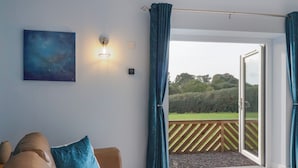 Ein Zimmer mit einem braunen Ledersofa, einem blauen Kissen, einer Wandleuchte und Blick auf einen Garten durch eine Glastür.