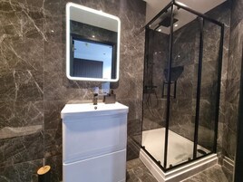 Un baño moderno con pared de mármol, un lavabo blanco, cabina de ducha de vidrio e iluminación integrada en un espejo.
