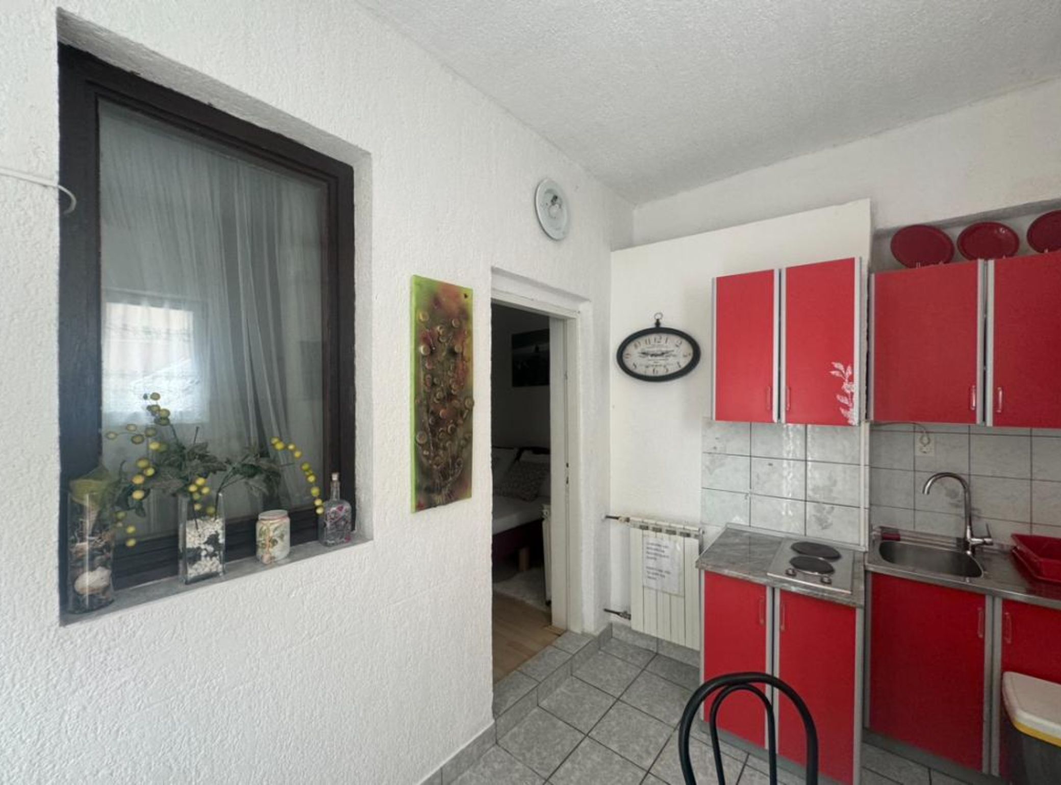 Une cuisine de taille réduite avec des placards rouges, une fenêtre ornée d’un rideau et une horloge au mur.