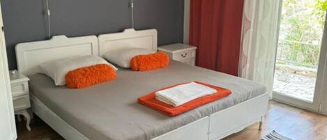 Ein Schlafzimmer mit Bett, Kissen, Nachttisch, Wanduhr und einem Fenster mit Vorhängen.
