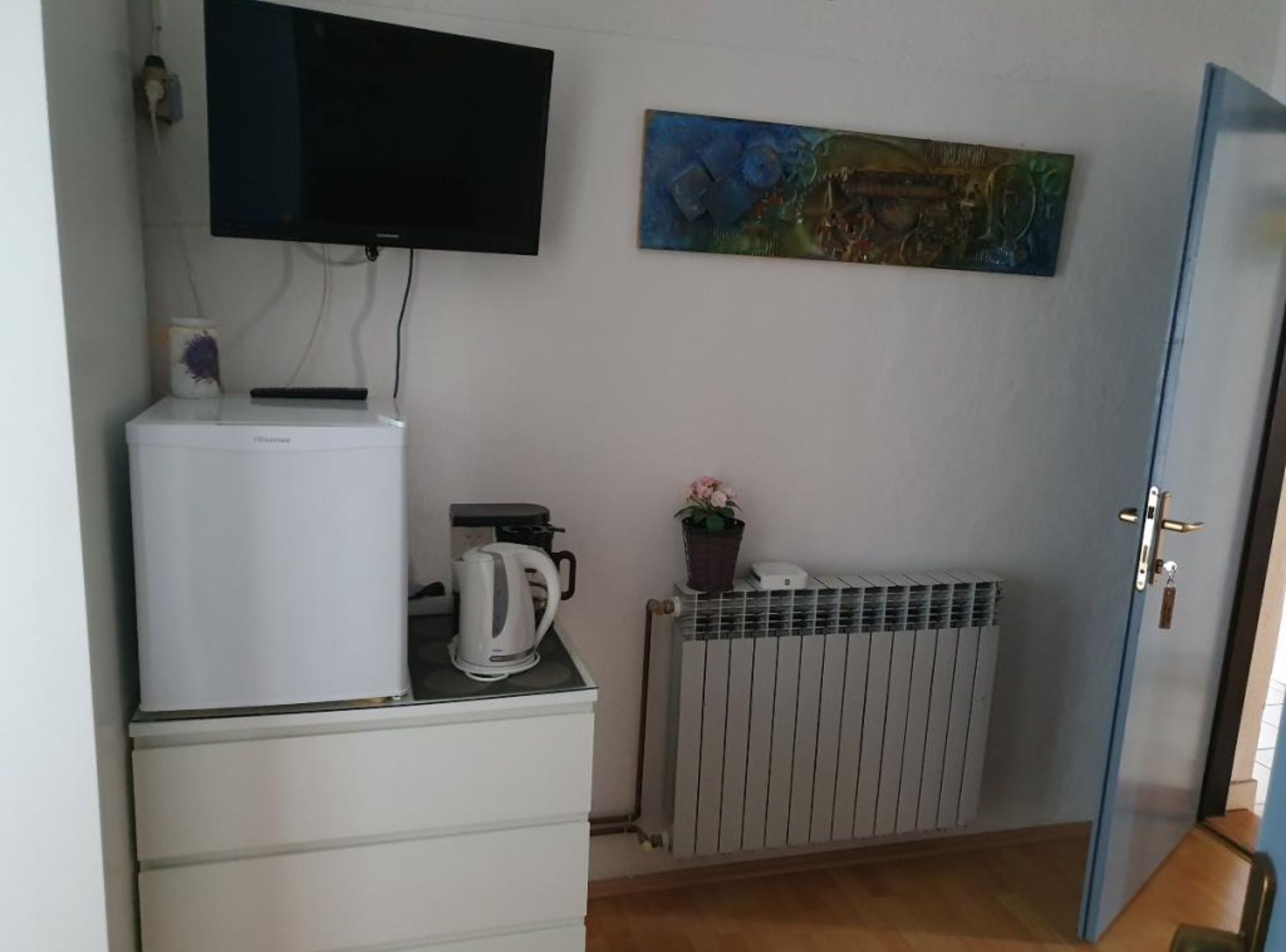 A room with a white refrigerator, a kettle, a radiator, and a television mounted on the wall.