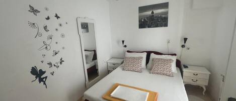 A bedroom with a bed, two bedside tables, a mirror, a wall clock, and a cityscape picture.