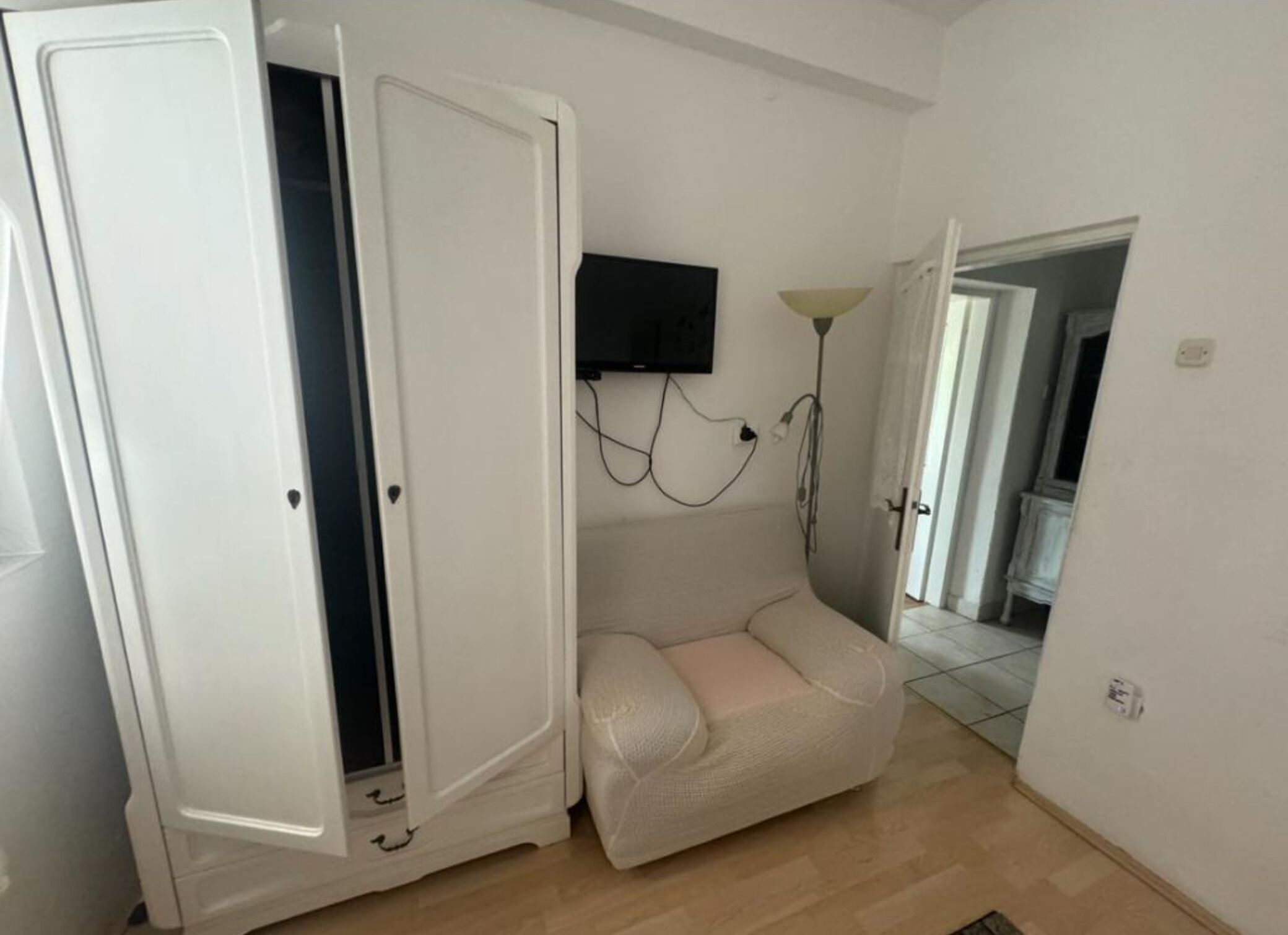 Une pièce avec une armoire blanche, un canapé beige, un téléviseur fixé au mur et une lampe.