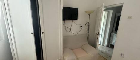 Une pièce avec une armoire blanche, un canapé beige, un téléviseur fixé au mur et une lampe.