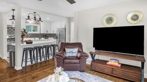 Een moderne keuken met witte kasten, een roestvrijstalen koelkast en een bar met krukken. Een bruine leren fauteuil met een kussen waarop 'BE OUR GUEST' staat, bevindt zich in de woonkamer, en een flatscreen televisie is bevestigd aan een houten standaard.