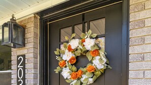 Een donkerbruine voordeur met een krans van witte en oranje bloemen. Een huisnummer '203' van baksteen is links zichtbaar.