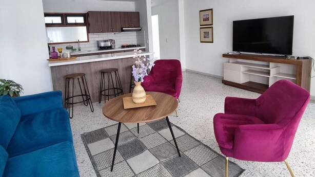 Una sala de estar moderna con una cocina al fondo, un televisor de pantalla plana y sillas coloridas.