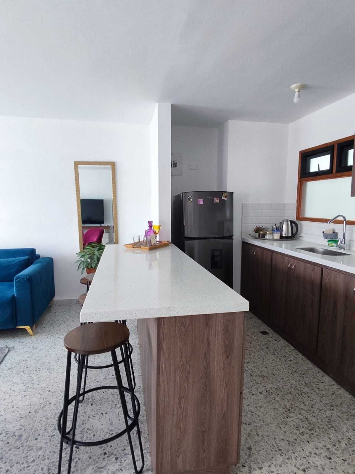 Une cuisine compacte dotée d’un îlot central, de meubles de rangement en bois foncé, d’un réfrigérateur noir et d’un canapé bleu dans l’espace de vie adjacent.
