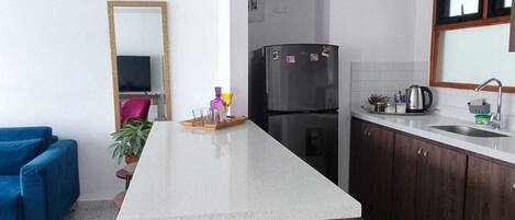 Une cuisine compacte dotée d’un îlot central, de meubles de rangement en bois foncé, d’un réfrigérateur noir et d’un canapé bleu dans l’espace de vie adjacent.