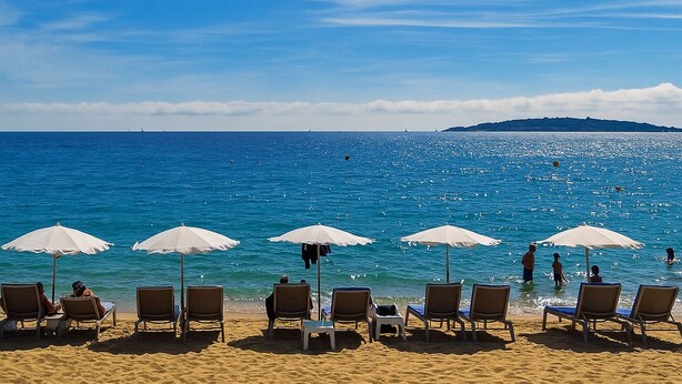 Une plage bordée de rangées de chaises longues et de parasols, une mer d’un bleu limpide et une île qui se dessine à l’horizon.