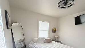 A bedroom with a bed, a standing fan, a window with blinds, a wall-mounted TV, and a small bedside table.