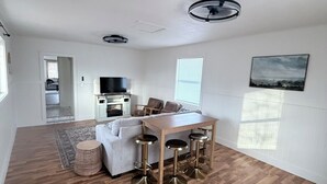 A spacious living area with a wooden floor, a sofa, a dining table with stools, a TV, and a kitchen in the background.