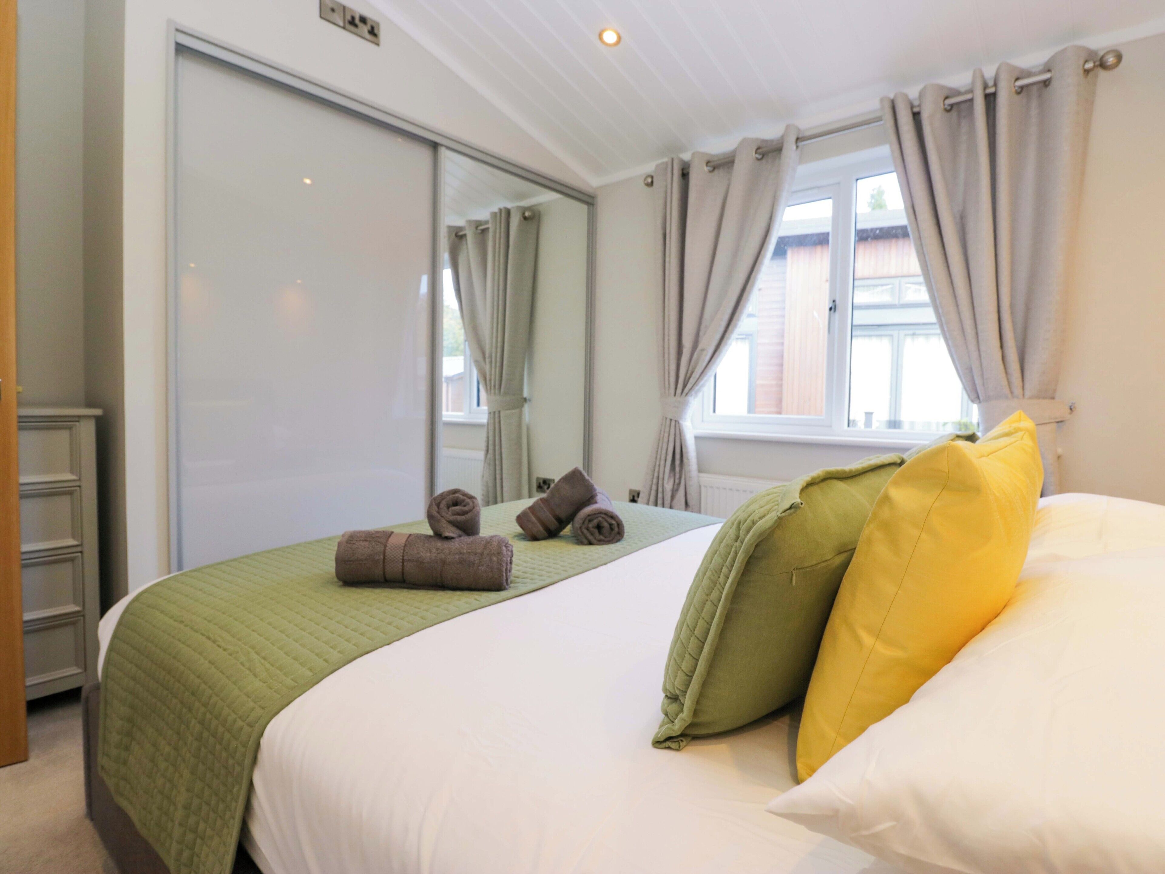 Una habitación de hotel con cama, una colcha verde acolchada, dos toallas enrolladas, un cojín verde y uno amarillo, una ventana con cortinas y un espejo.