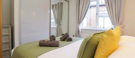 Una habitación de hotel con cama, una colcha verde acolchada, dos toallas enrolladas, un cojín verde y uno amarillo, una ventana con cortinas y un espejo.
