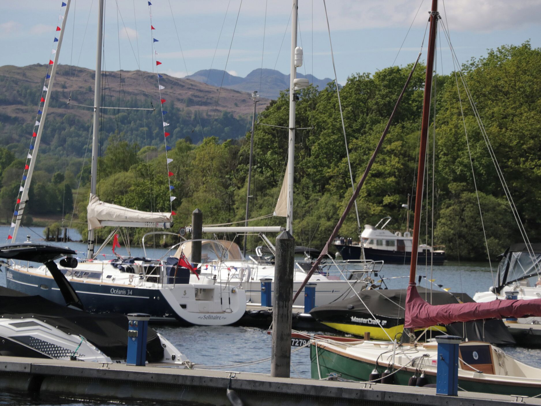 A marina with various sailboats docked, surrounded by lush greenery and hills in the background.