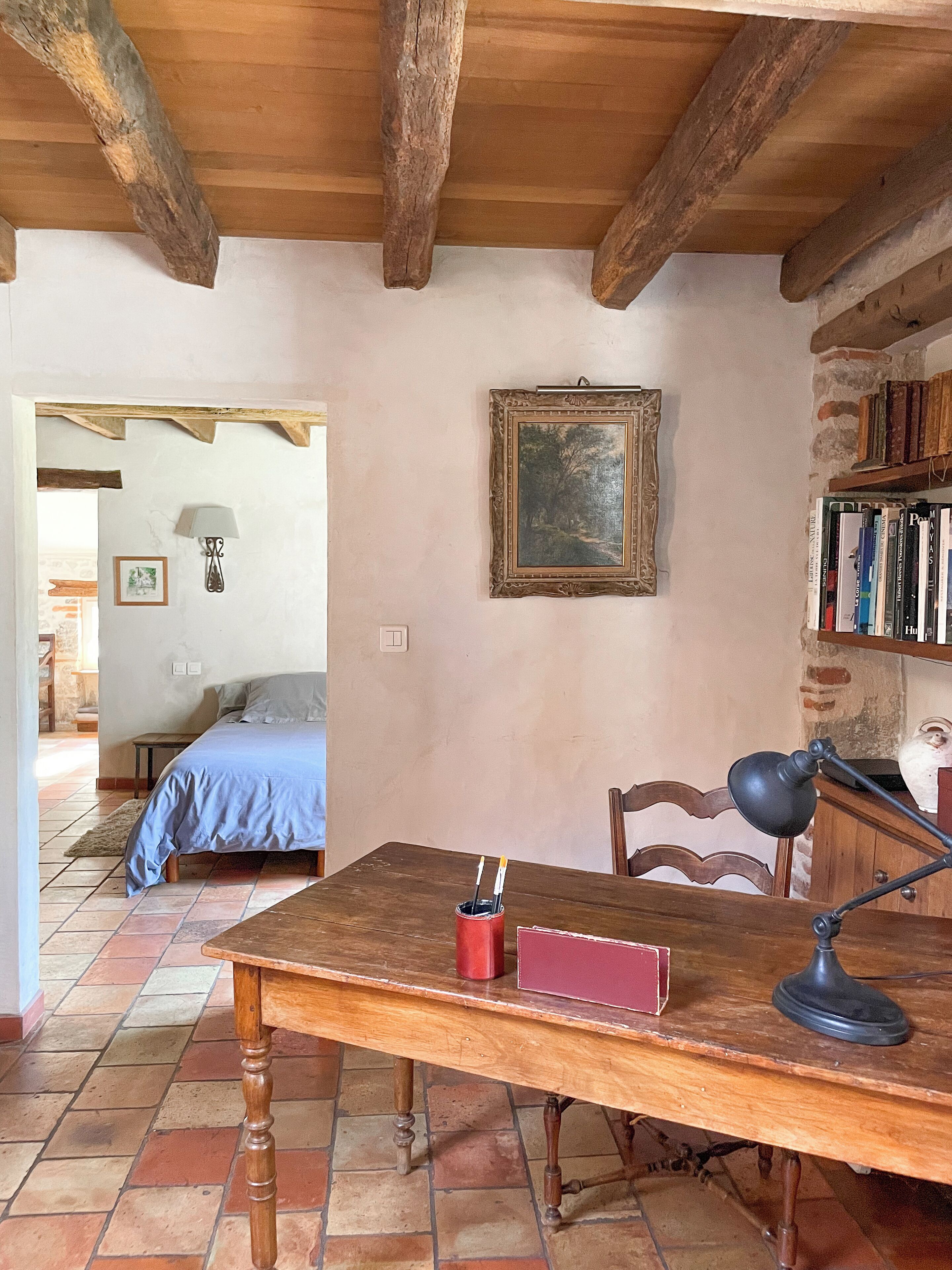A rustic room with a wooden table, a chair, a desk with a lamp, a bookshelf, and a bed in the background.