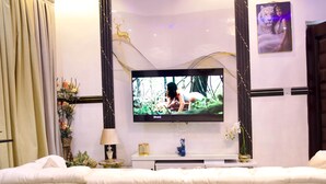 A modern hotel room with a large flat-screen TV mounted on the wall, a bed with white bedding, a small side table with a lamp, and decorative elements including a framed picture of a tiger and a golden deer sculpture. - ftrealtyproperties (Accra)