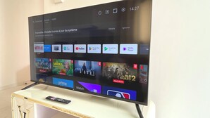 Un televisor mostrando la interfaz de un servicio de transmisión con varios iconos de aplicaciones y un control remoto sobre un soporte.