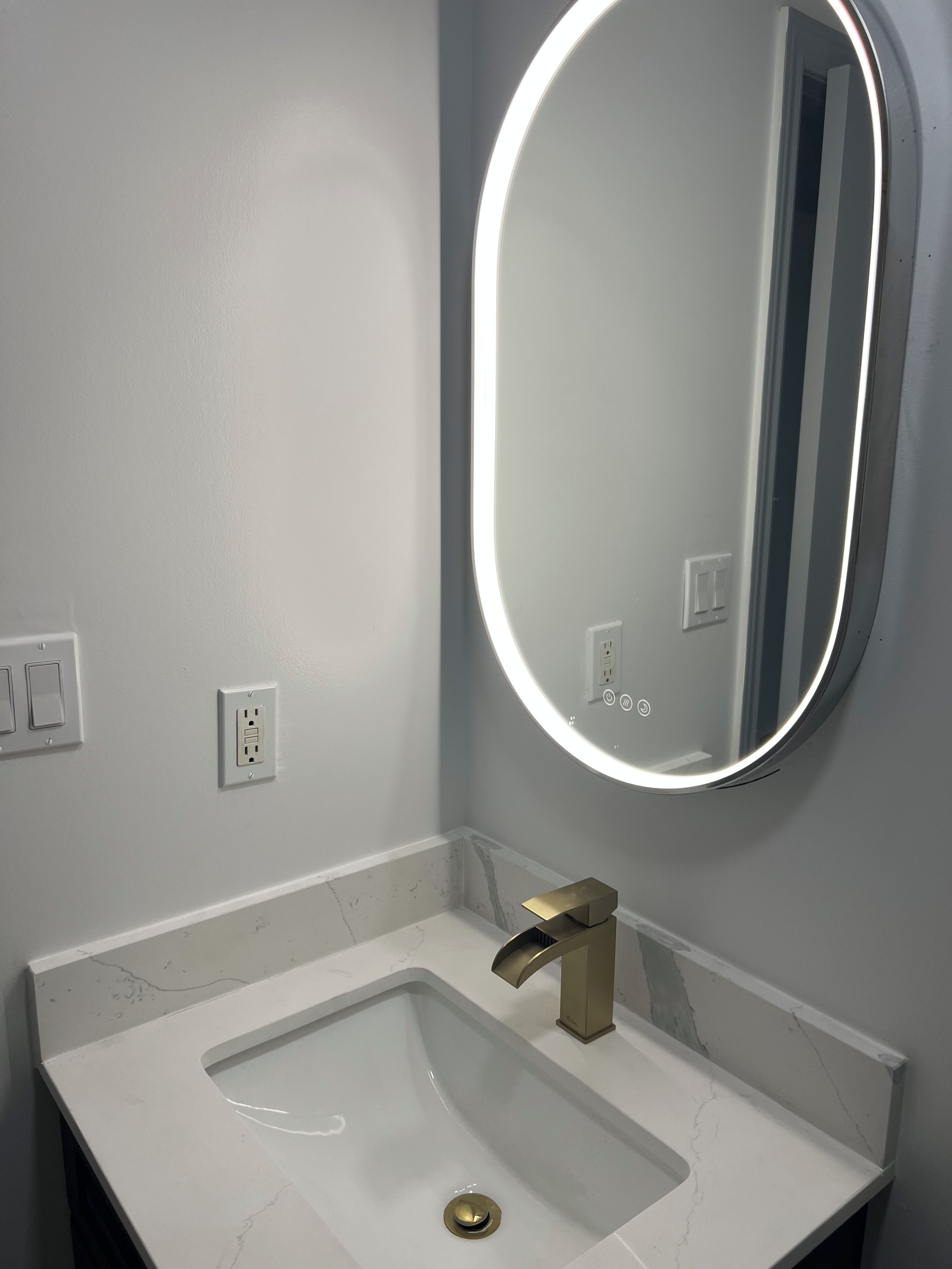 A bathroom with a white sink, a gold faucet, and a wall-mounted mirror with a light.