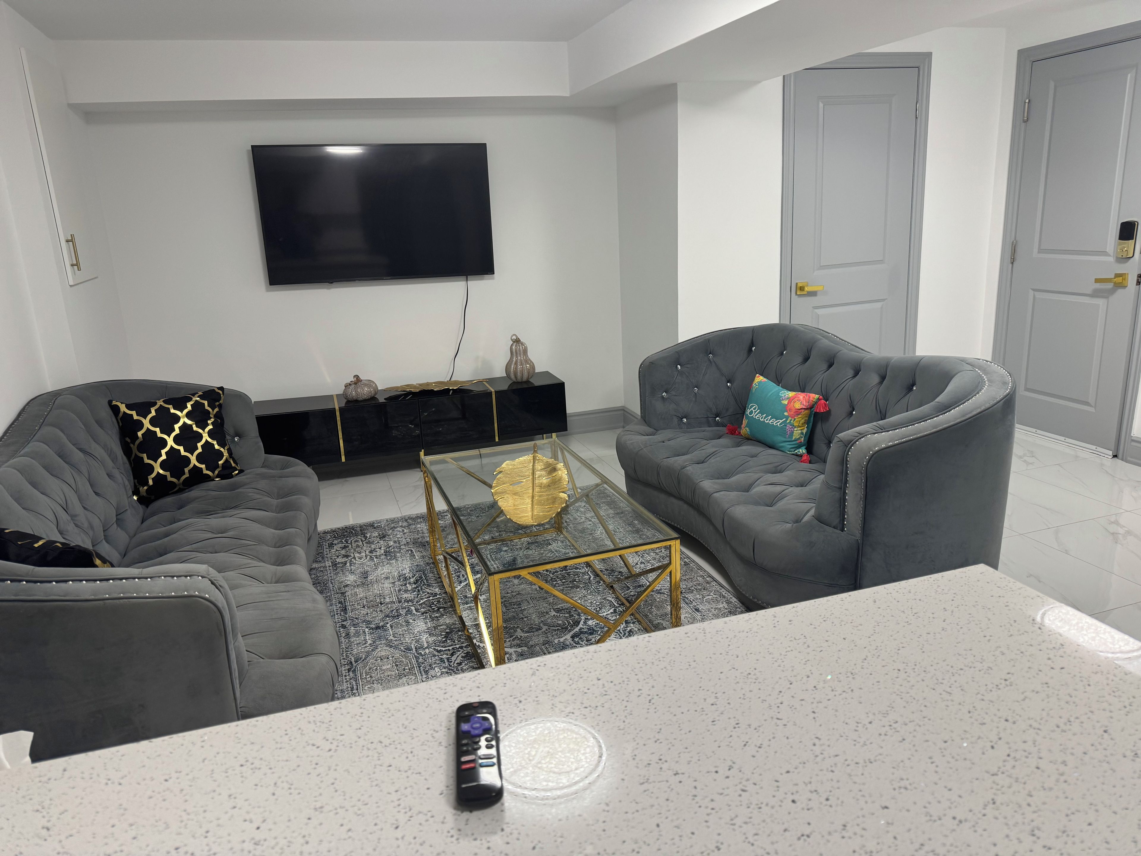 A modern living room with a flat-screen TV, two grey sofas, a coffee table, and a door with a keypad lock.