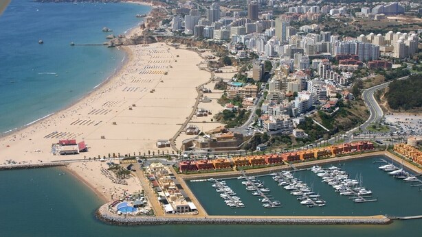 Vue aérienne d’une ville côtière avec une plage, un port de plaisance et une zone urbaine densément peuplée.