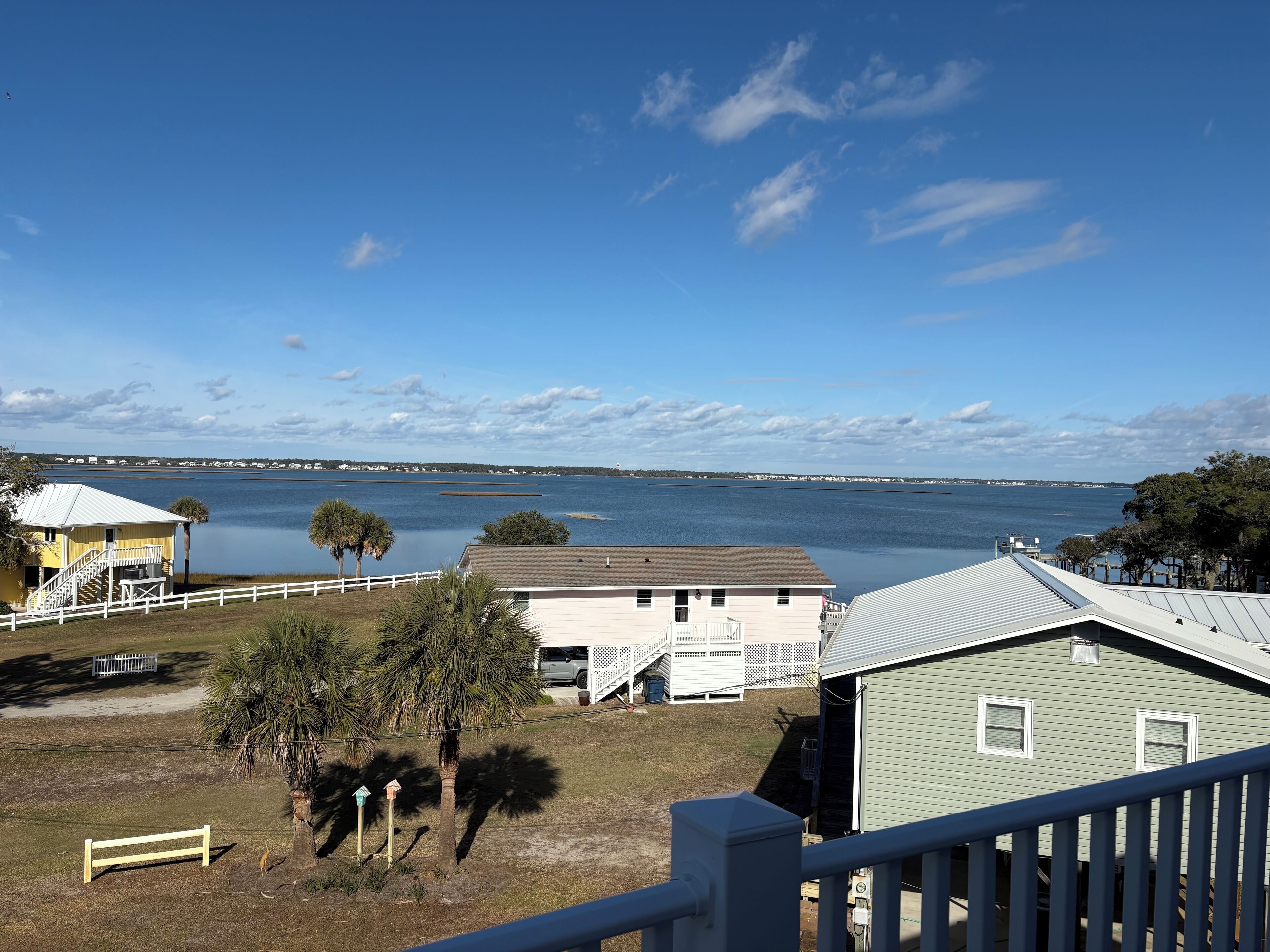 A coastal view with a clear sky, a calm body of water, and a row of houses with a white fence and palm trees.