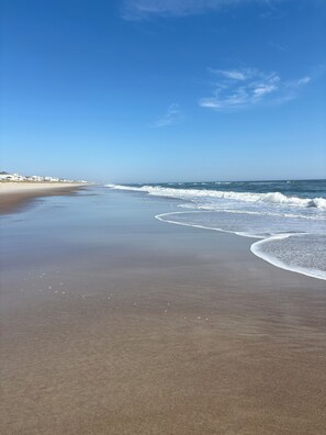 A beach with a clear sky, waves, and a few buildings in the distance.