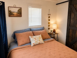 Un lit avec une couvre-lit à motifs, deux oreillers, une table de chevet avec une lampe, une fenêtre avec des stores et un tableau accroché au mur.