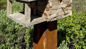 A rustic wooden birdhouse with a slanted roof, set against a backdrop of a clear blue sky, green foliage, and distant snow-capped mountains. - Da Hotel OCt (North Vancouver)