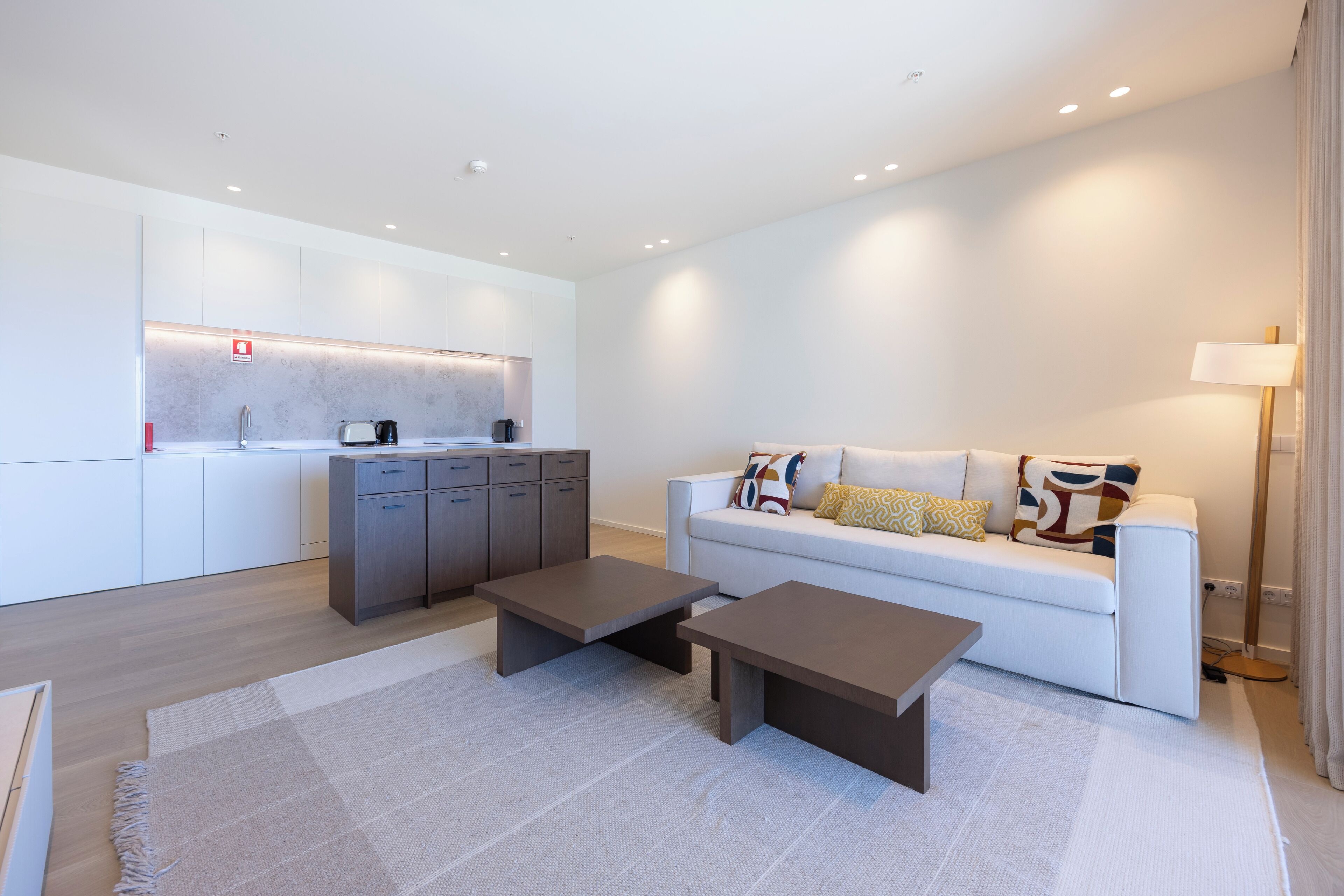 A modern living room with a sofa, two coffee tables, a kitchen area with cabinets, and a lamp.