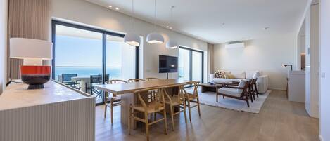 A modern hotel room with a large dining table, wooden chairs, a sofa, and a TV.