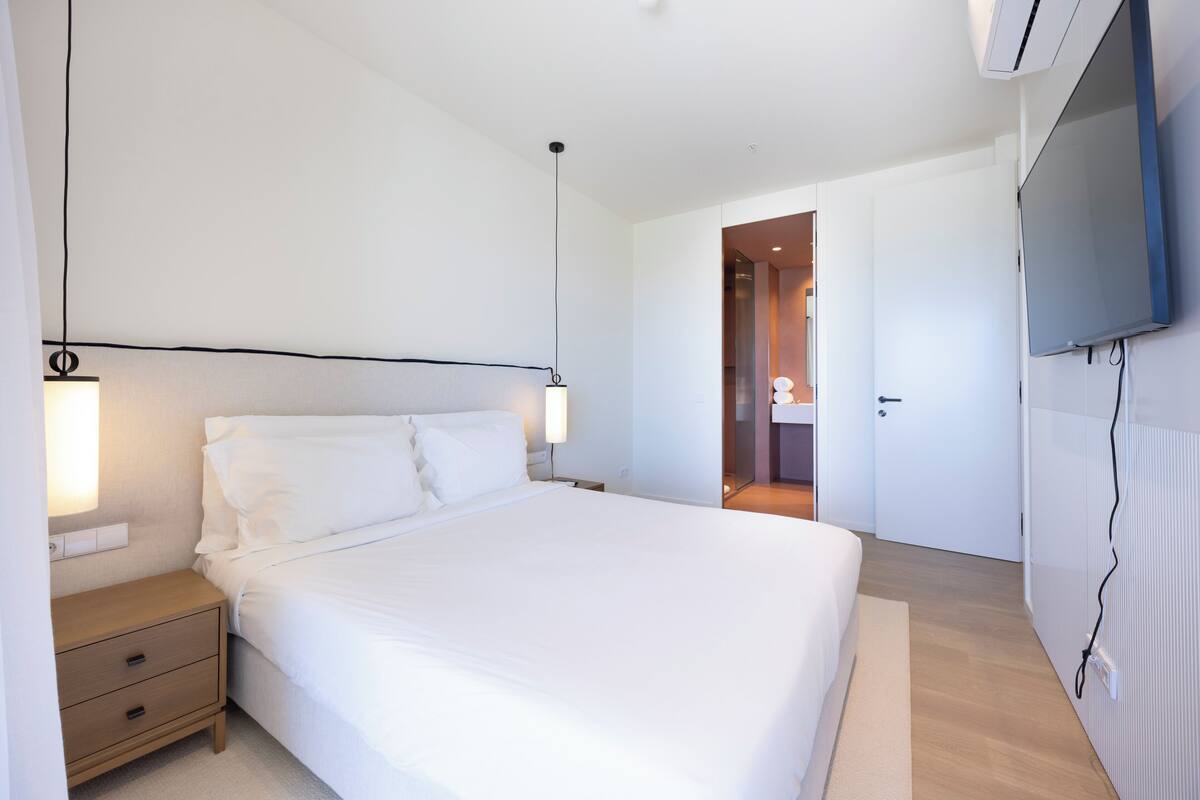 A modern hotel room with a large bed, bedside tables, a TV, and a door leading to a bathroom.
