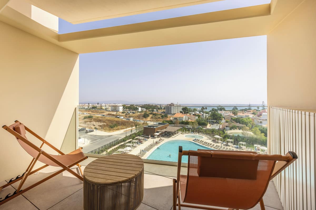 A balcony with two lounge chairs, a small round table, and a view of a swimming pool and buildings.