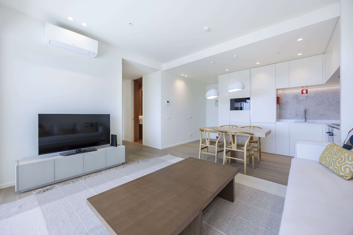 A modern living room with a flat-screen TV, a wooden coffee table, and a dining area with a small kitchen in the background.