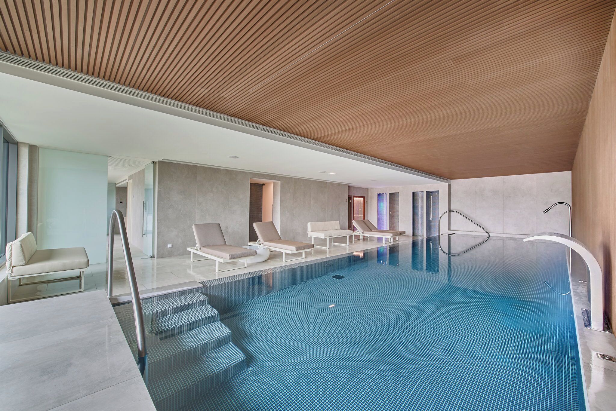 A modern indoor swimming pool with clear blue water, lounge chairs, and a wooden ceiling.