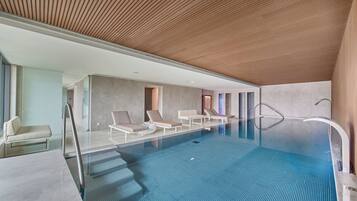 A modern indoor swimming pool with clear blue water, lounge chairs, and a wooden ceiling.