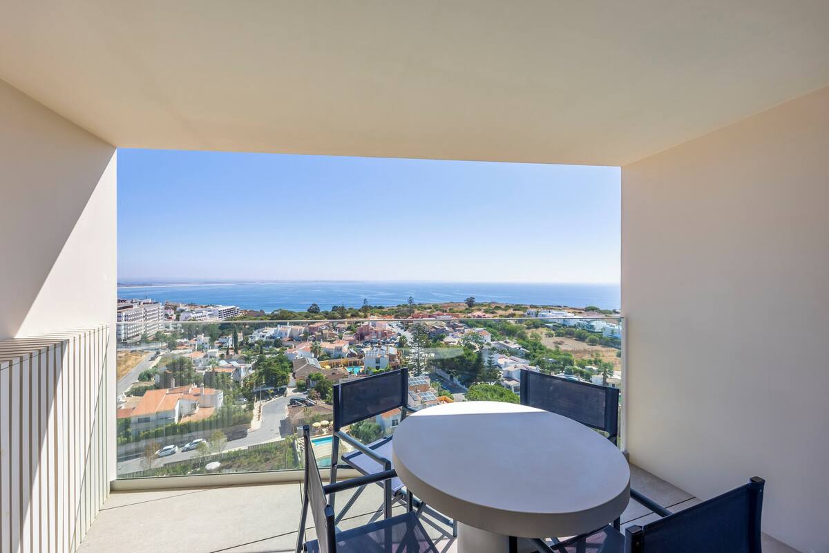 A balcony with a round table and chairs, overlooking a coastal town with buildings and a clear sky.