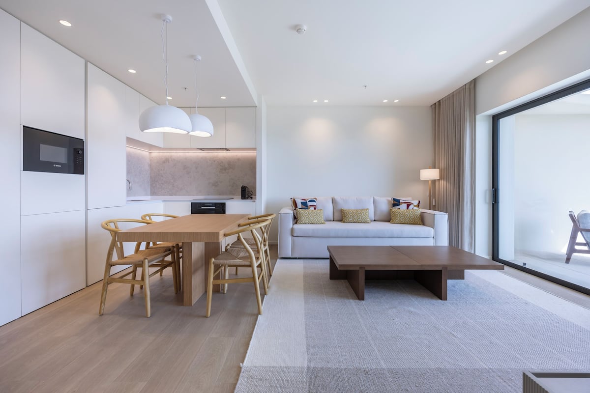 A modern living area with a dining table, chairs, a sofa, and a coffee table. There is a kitchen area with a microwave and a hanging light fixture.