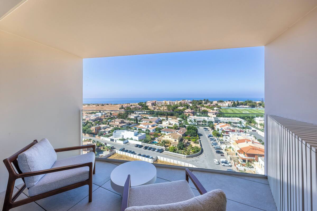A balcony with two chairs and a small table, overlooking a residential area with a clear view of the sea.