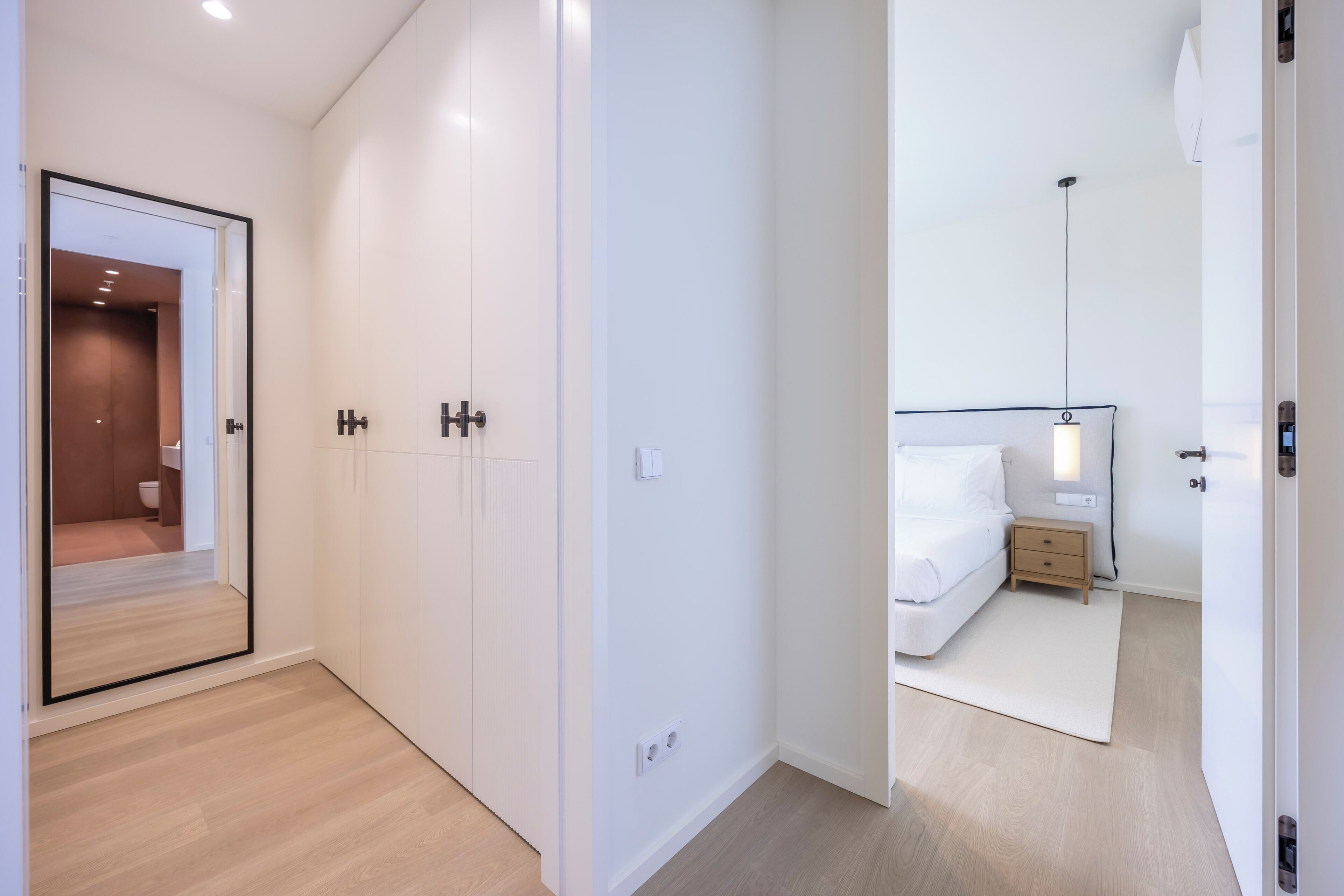 A modern hotel room with a large wardrobe, a bedside table with a lamp, and a view into the bathroom.