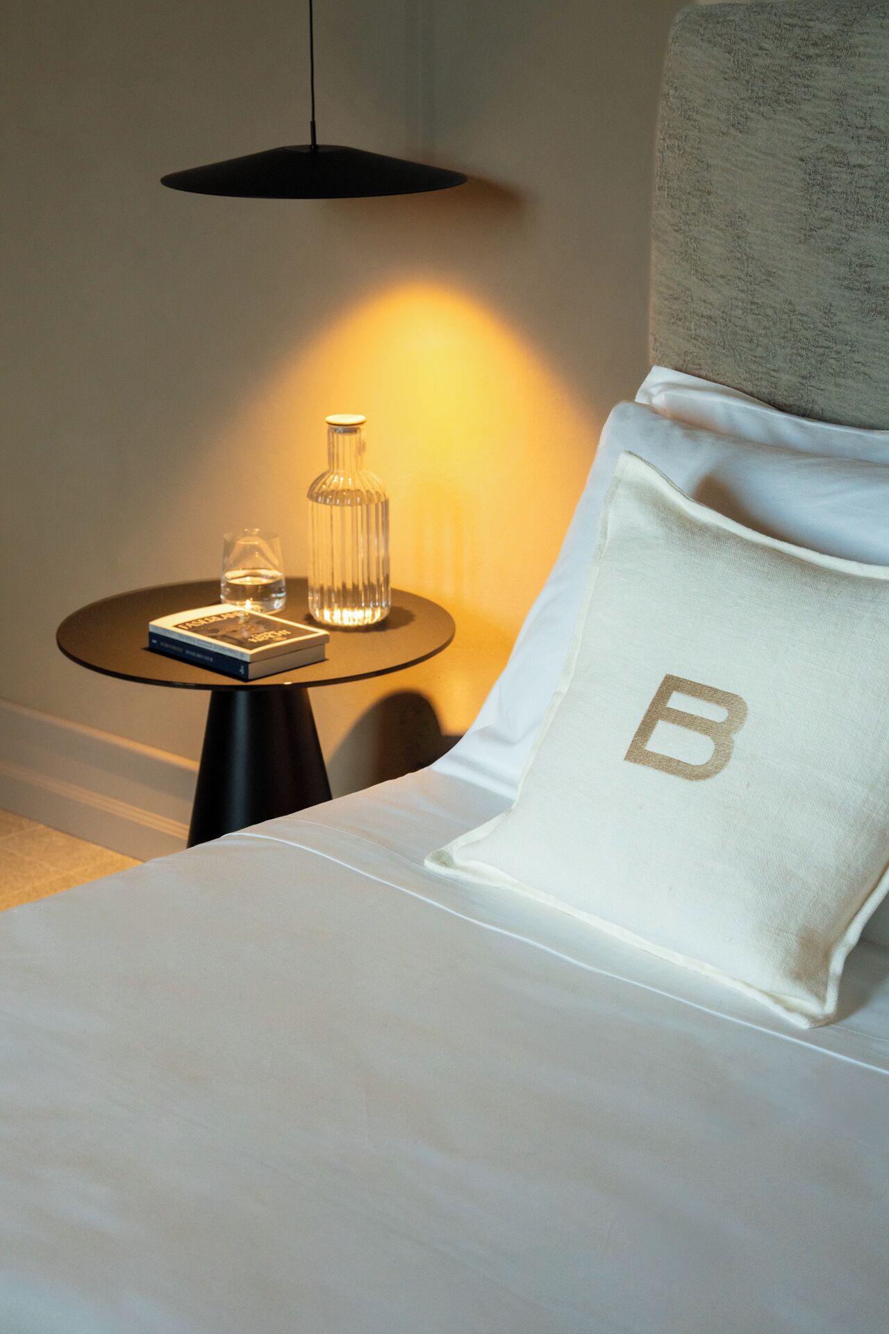 Uma cama bem arrumada com um travesseiro marcado com uma letra 'B' grande, uma mesa de cabeceira com um livro e uma garrafa de vidro, e um abajur projetando um brilho quente.