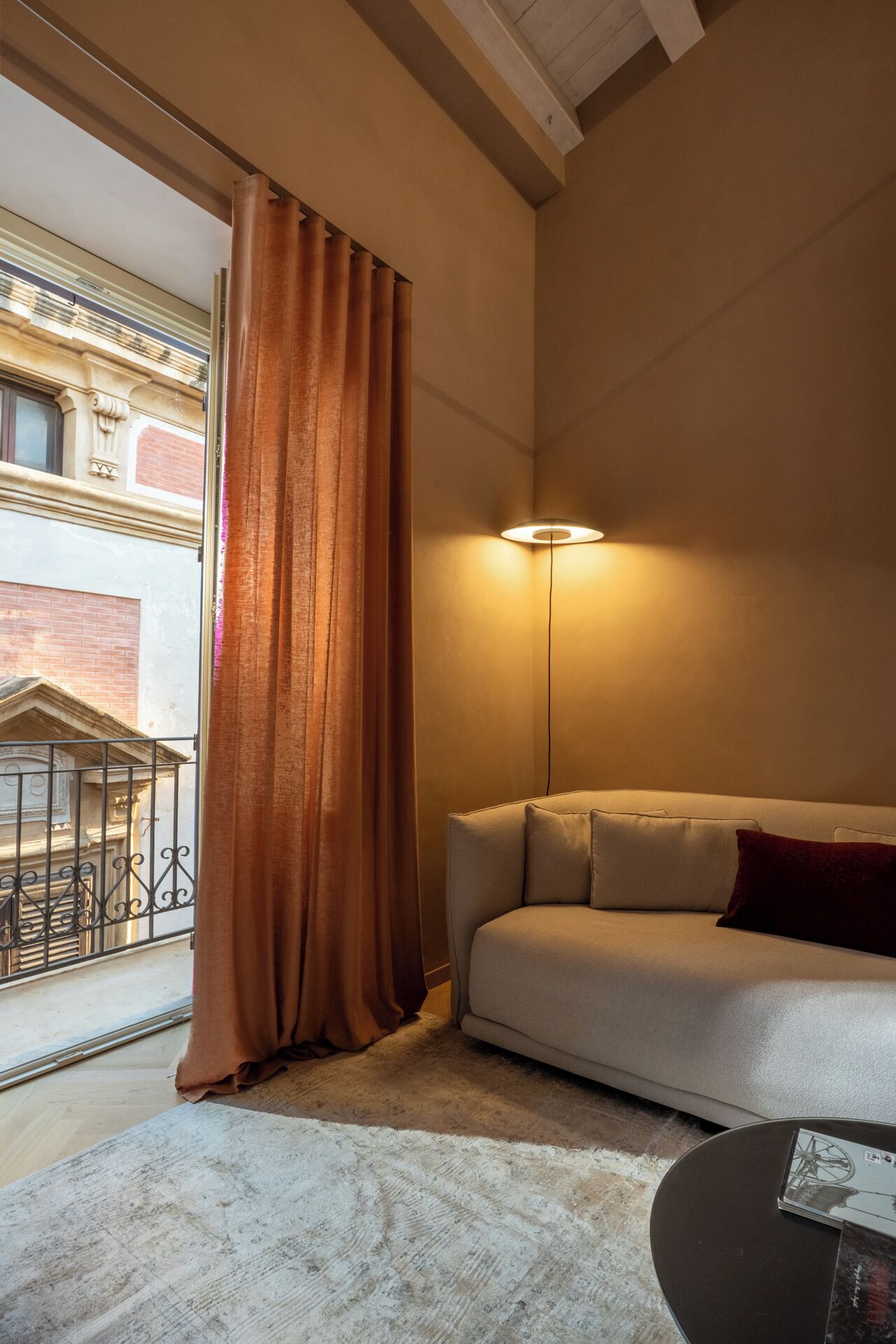 A room with a sofa, a lamp, a window with curtains, and a view of a building outside.
