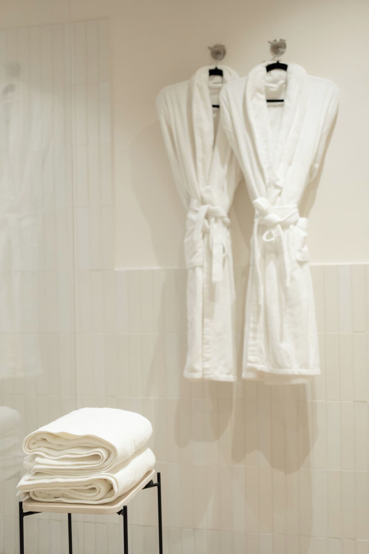 A bathroom with two white robes hanging on hooks and a stack of folded white towels on a small table.