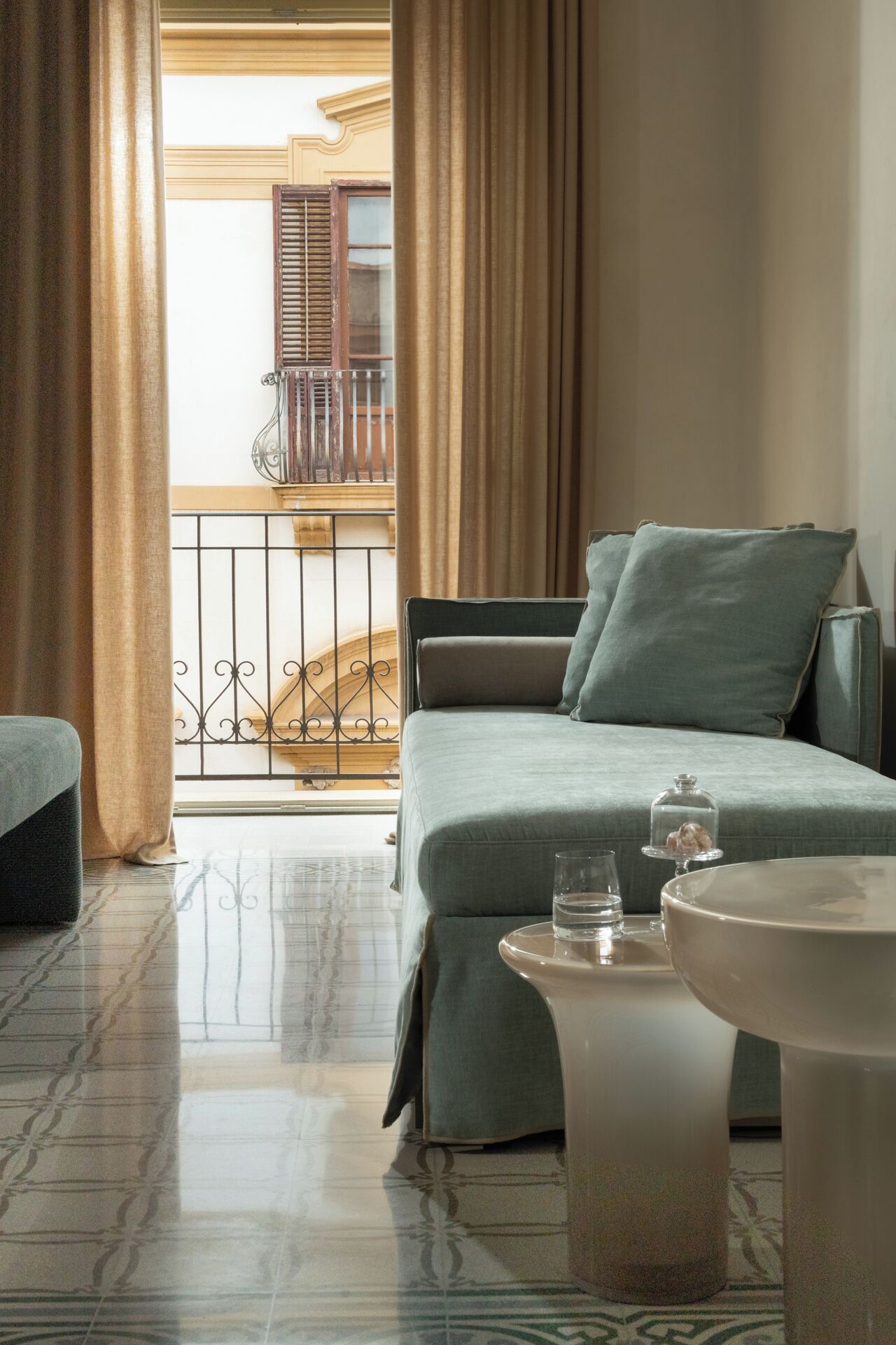 A hotel room with a bed, a small table with a glass and a decorative item, and a view of a balcony with a railing and a window with shutters.