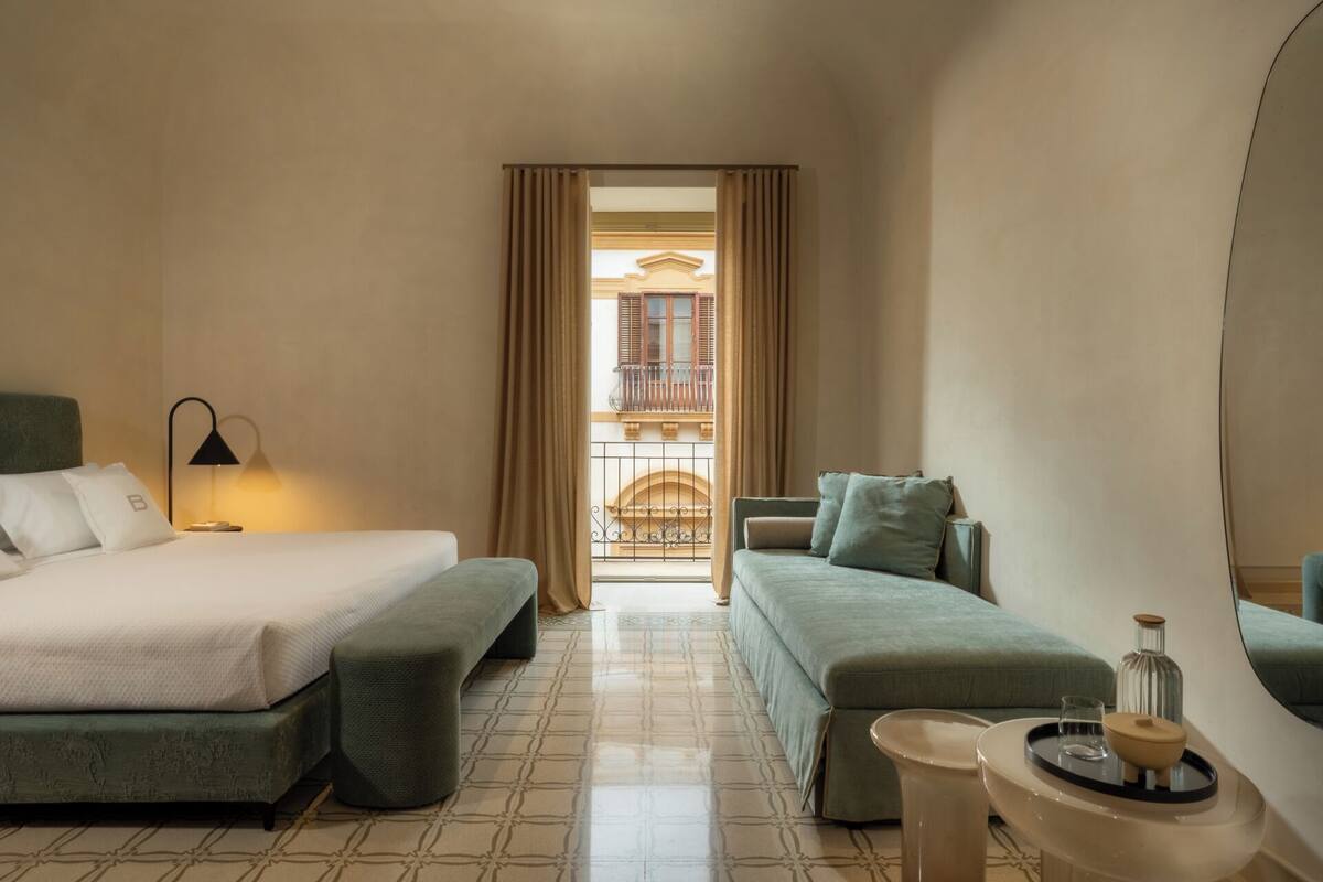Une chambre d’hôtel avec un lit, un canapé, une petite table avec une bouteille et un verre, et une vue sur un balcon doté d’une balustrade.