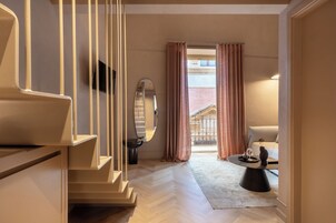 Quarto de hotel moderno com escada de madeira, um espelho grande, área de estar com mesa e vista para um edifício através de uma porta de vidro.