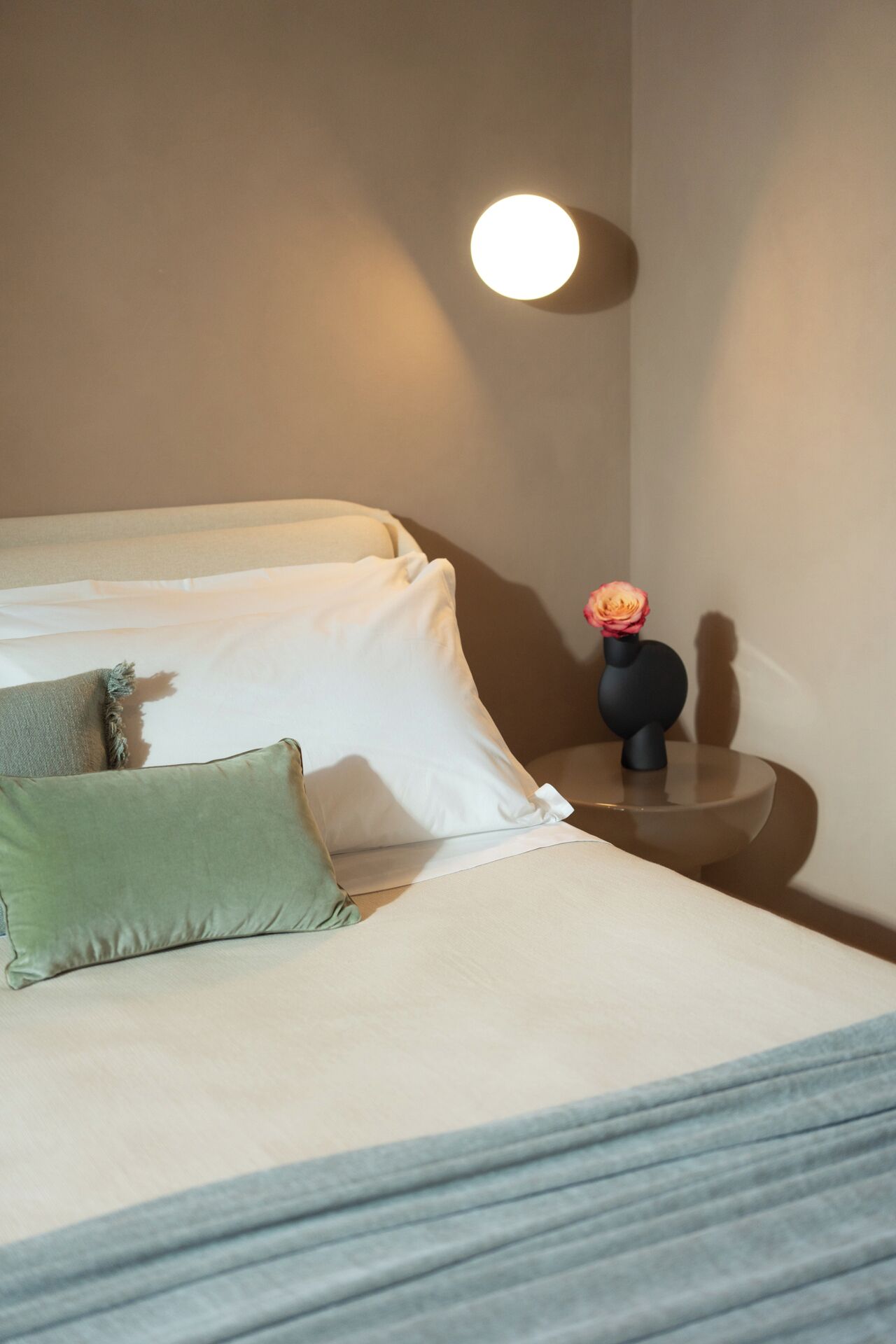 Un lit bien fait, avec des oreillers, une table de chevet ornée d’un vase de fleurs et une lampe fixée au mur.