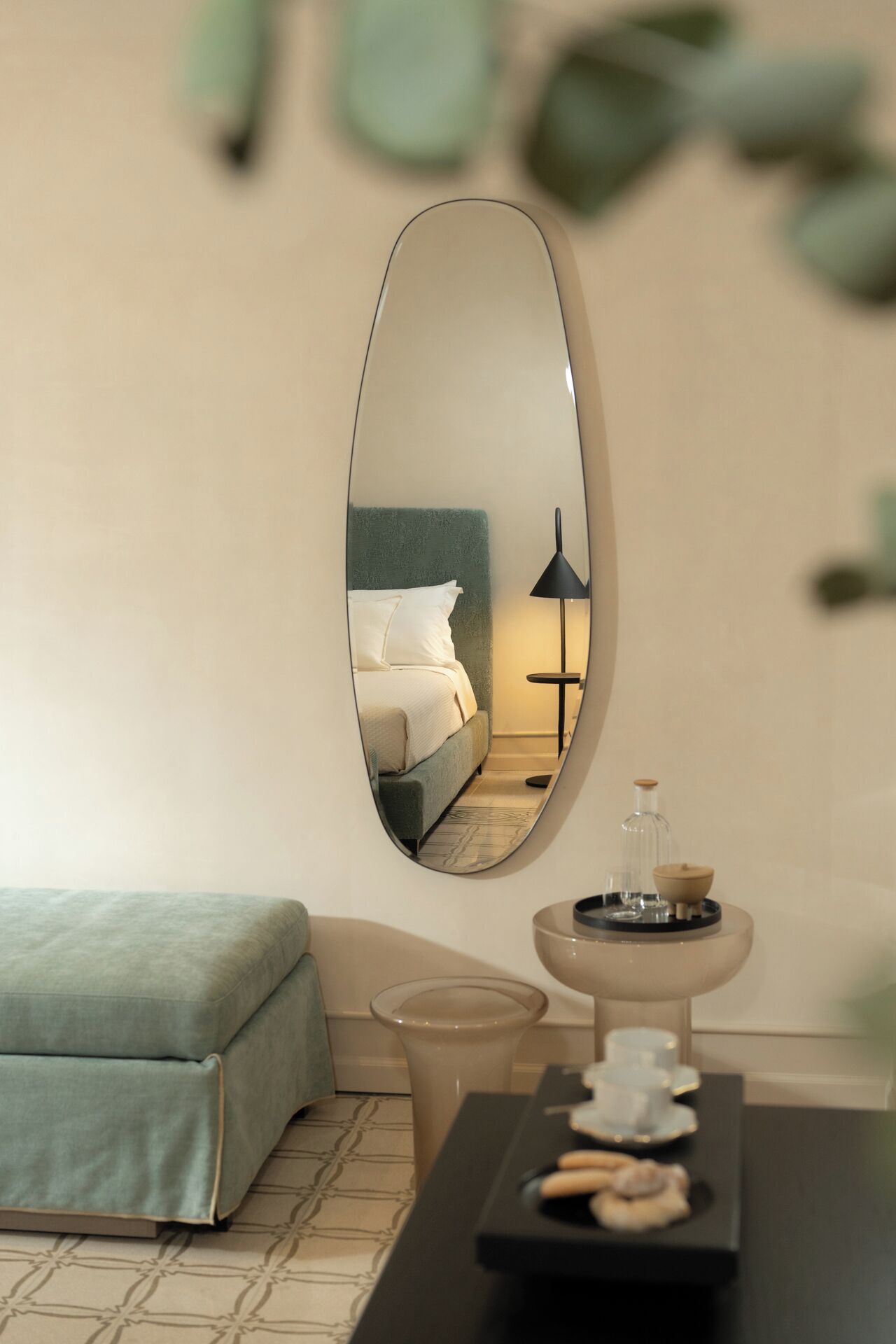 A modern hotel room with a large oval mirror reflecting a neatly made bed, a bedside lamp, and a small table with a tray of cookies.