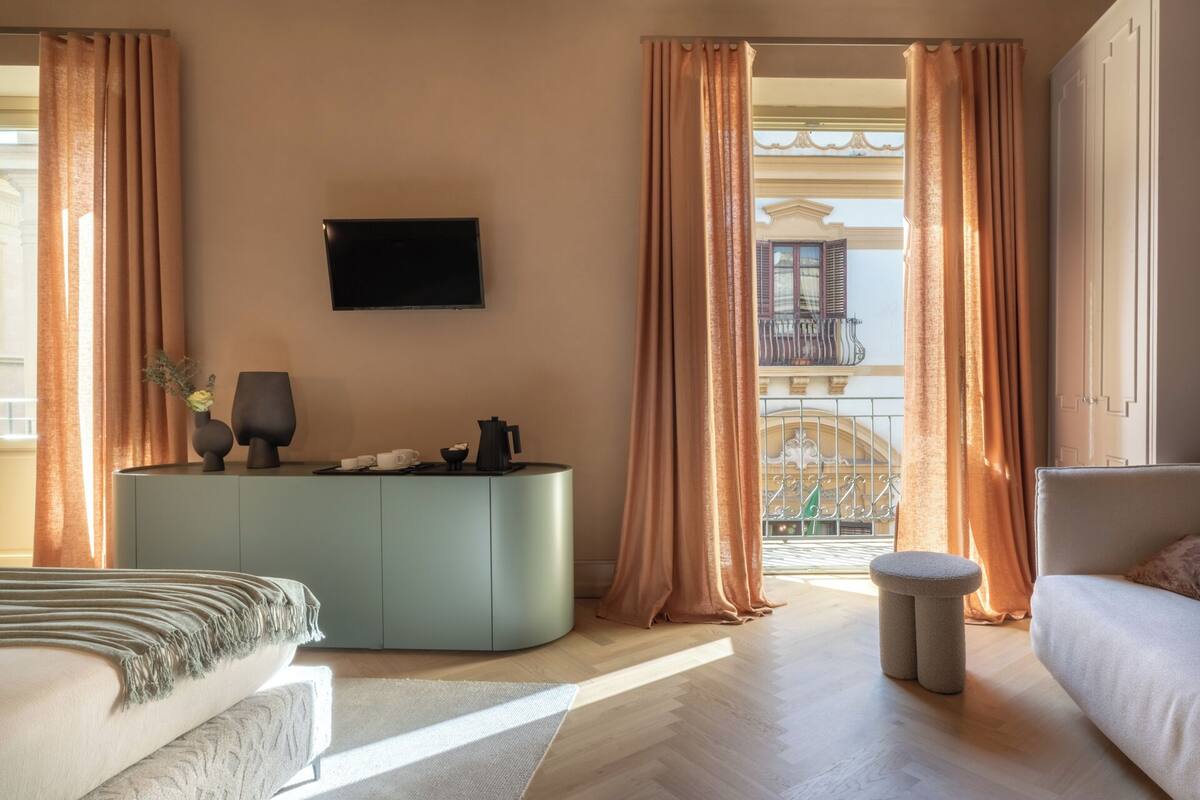 Une chambre d’hôtel moderne dotée d’un grand lit, d’un coin salon, d’une télévision à écran plat et offrant une vue sur un balcon avec un bâtiment en arrière-plan.