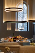 Un buffet per la colazione con diversi dolci e frutta, ambientato in una stanza con ampie finestre e lampade pendenti moderne.
